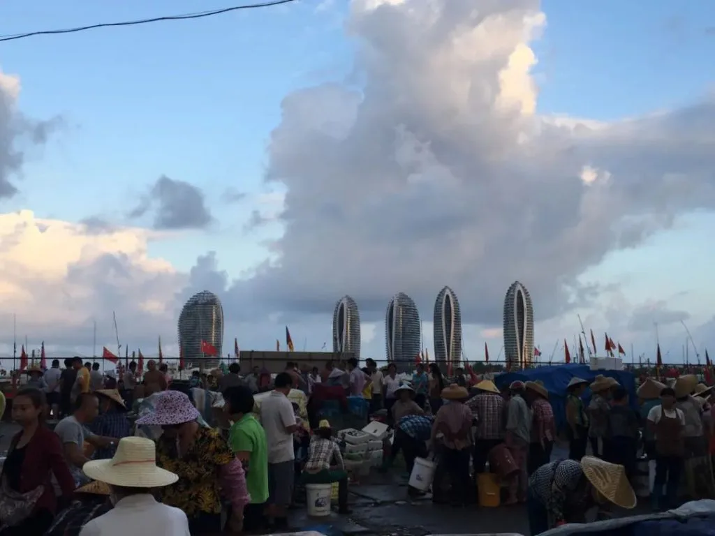 码头上人头攒动,多数人戴着草帽、旁边放着塑料桶忙碌地进行买卖;远处天际线是造型独特的螺旋状高层建筑,即那家五星级酒店,天空布满层叠的云团。