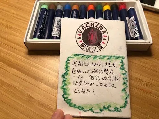 手举着zine封底,上方印着“VaChina 阴道之道”圆形徽标,四周描绿色波浪蜡笔边框,桌上放着一盒蜡笔。