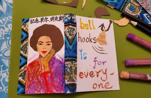左页是bell hooks的彩色插画肖像(爆炸头、彩色斗篷),上方写着“纪念贝尔·胡克斯”,镶蓝色花纹边框;右页手写文字旁挂着写有“Live Freedom”的小标签,周围散落彩色蜡笔和碎纸。