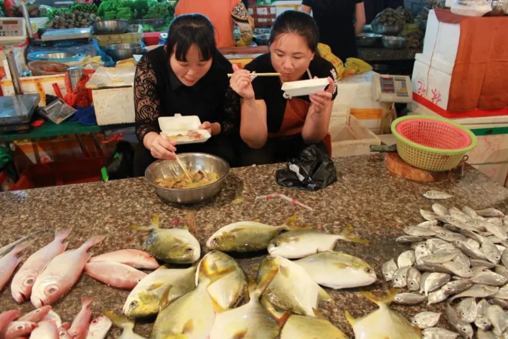 两位女摊主坐在鱼摊后,用外卖饭盒就着快餐吃饭,面前的摊位台面上摆满了金鲳鱼、鲷鱼等多种鲜鱼。