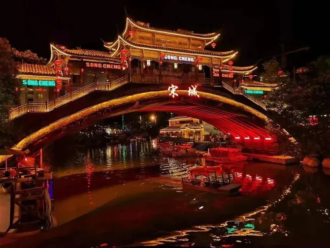 金色拱桥上悬挂“宋城”红色大字与“SONG CHENG”霓虹招牌,飞檐楼阁灯火通明,桥下河面停靠着红色游船,灯光倒映水中。