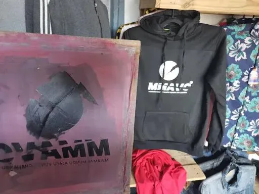 左侧是木框丝网印花模具,印有反向的图案与文字;右侧挂着一件印有品牌标志的黑色连帽衫,旁边还有红色、蓝色等待加工或售卖的衣物。
