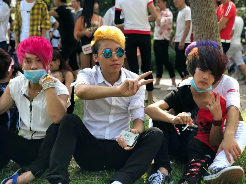 三名染着粉、黄、紫色高耸发型的青年坐在草地上,中间一人戴墨镜比出'V'字手势,两侧的人戴着口罩。