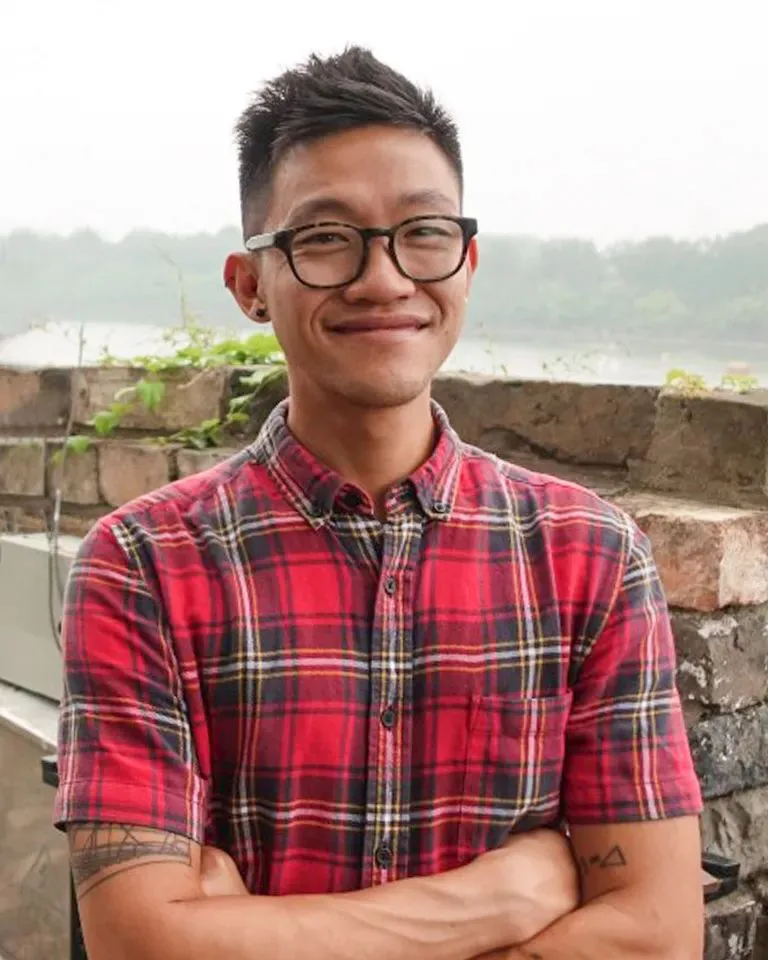 徐庄骅戴黑框眼镜，穿红黑格纹衬衫，双臂交叠，微笑着站在河边石墙前。