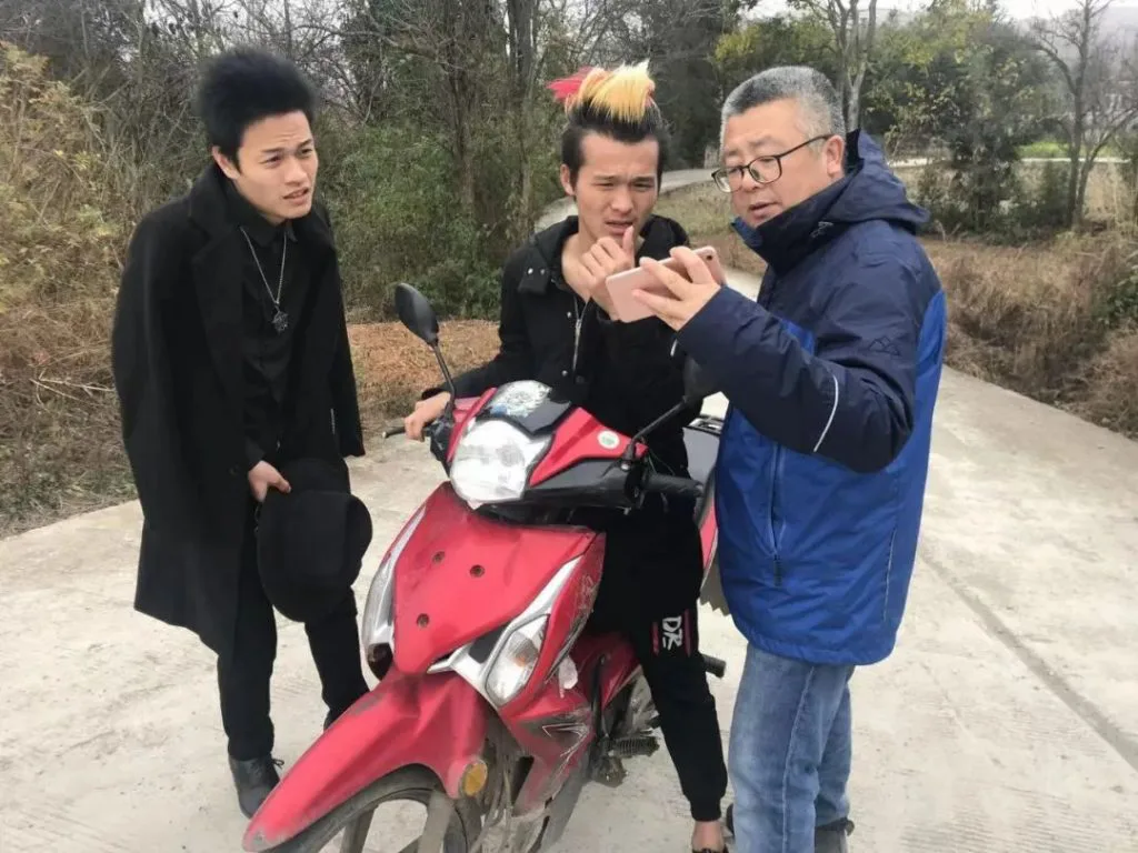 导演李一凡与两名留着夸张竖发的青年站在一辆红色摩托车旁的乡间小路上,其中一人正给他看手机。