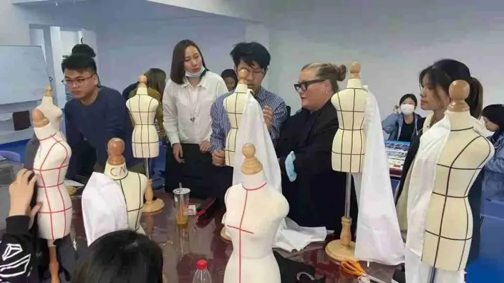 一位外教在服装设计课上示范人台立裁,多名学生围在摆满小型人体模型的桌前观看。