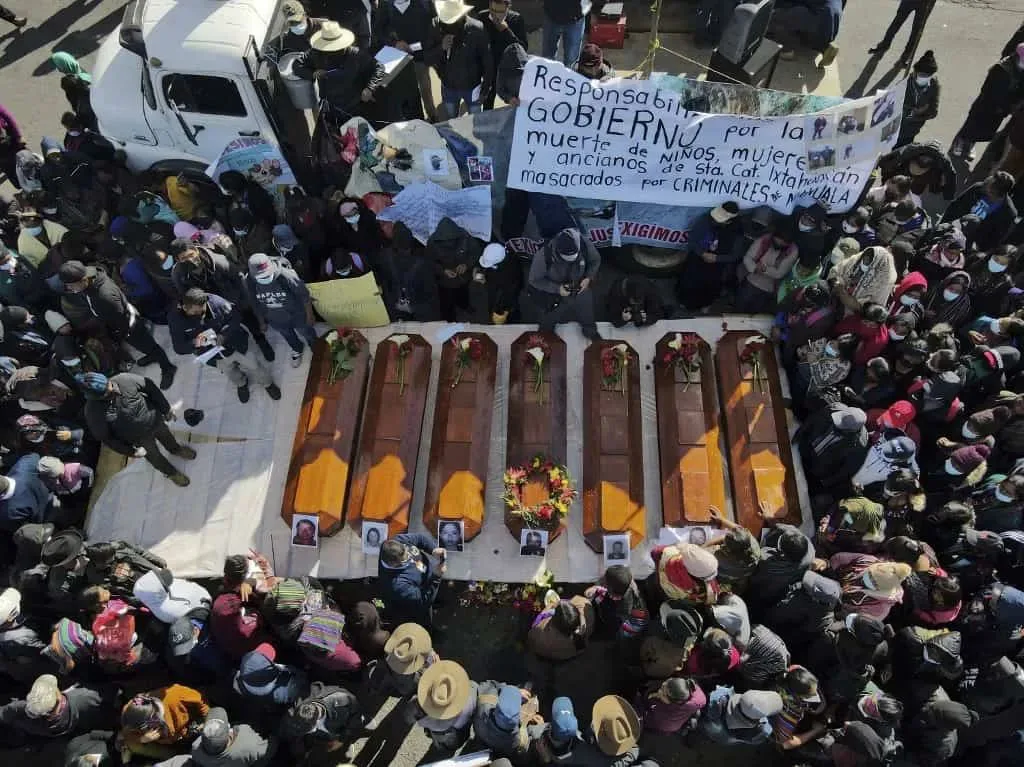 俯瞰视角，七具棺材整齐排列在地面，棺盖上放着遇难者照片和花束，周围站满悼念民众，背景高举一幅西班牙语横幅，谴责政府对儿童、妇女和老人遭黑帮屠杀负有责任。