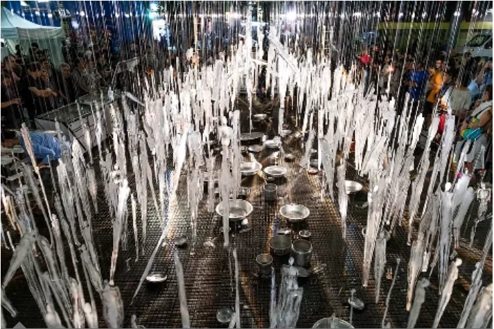 夜晚的广场上，成排细长的冰质人形雕塑悬垂而下，下方铁网地面摆着接融水的金属托盘，两侧站满围观人群。