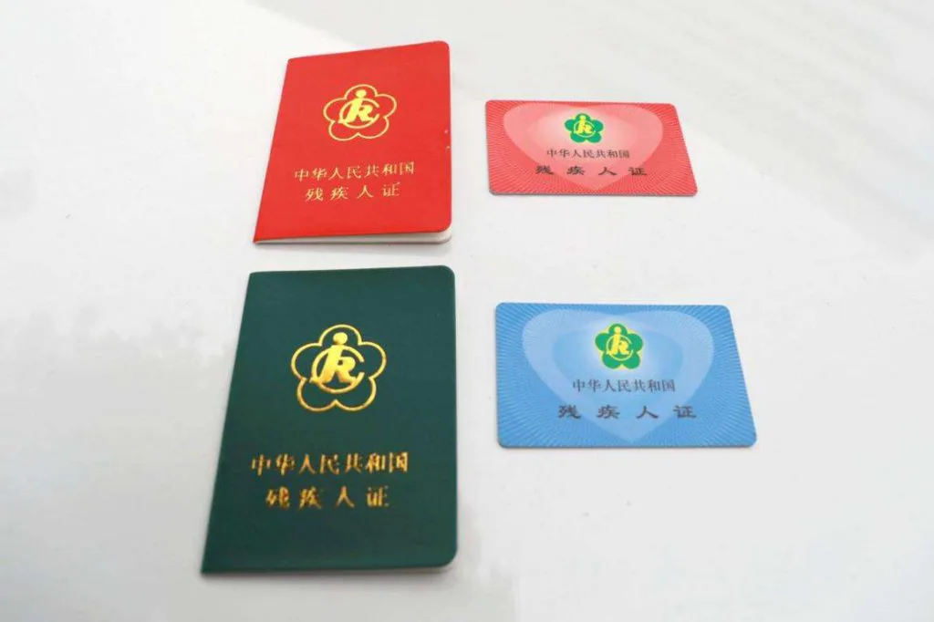 两种版式的残疾人证并排摆放：红色和深绿色的证件本，以及粉色和蓝色的卡片式证件，均印有同一朵小红花徽标。