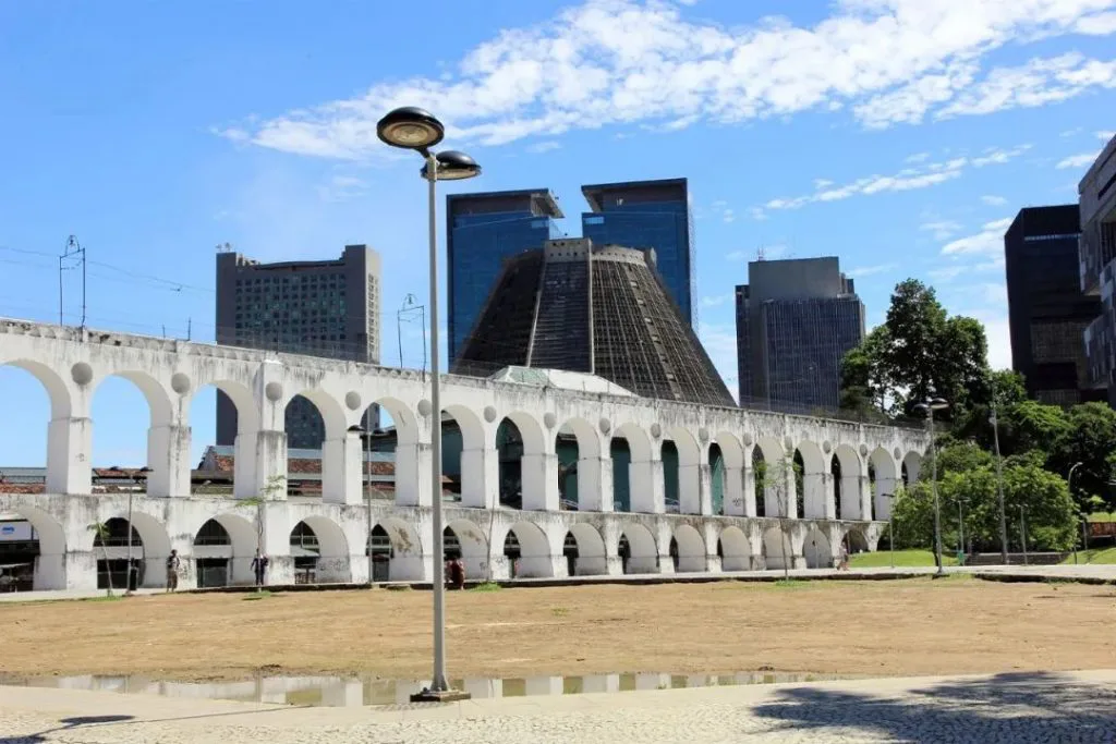 白色双层连拱建筑绵延于开阔广场旁，背景是里约现代高楼和圆锥形的都主教座堂，前景地面有积水倒影。