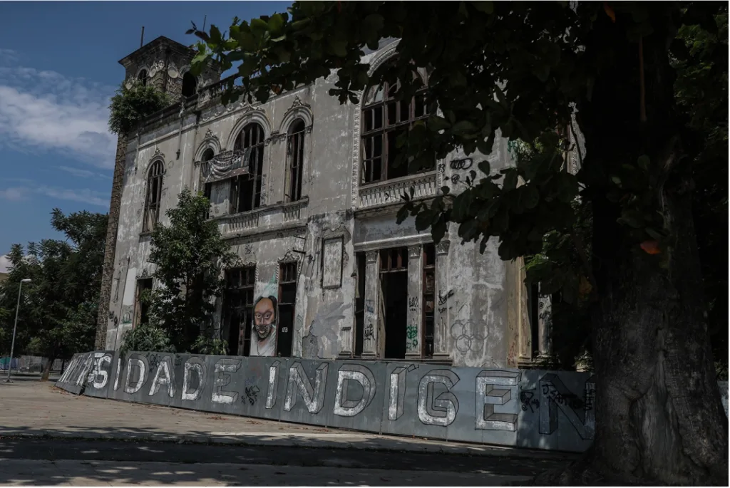 一栋门窗残破、墙面斑驳的老建筑，外墙涂鸦，墙根立着写有'…SIDADE INDÍGENA'（原住民大学）字样的招牌，墙上还有一幅人像涂鸦。