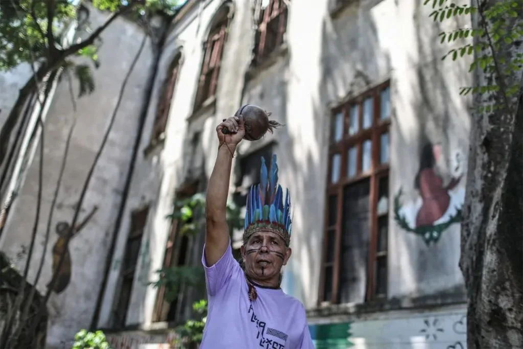 一位脸绘图腾、头戴蓝色羽毛头饰的男性举起手中的沙锤，身后老建筑墙上绘有原住民人物壁画。
