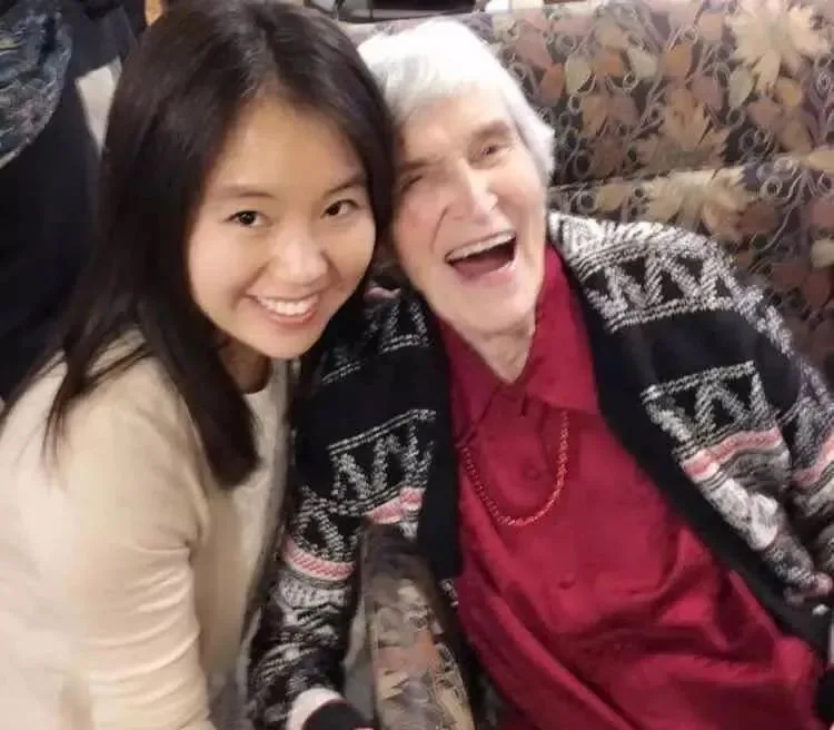 两人靠坐在花纹沙发上开怀大笑自拍,年长的白发女性穿黑白提花外套配红色上衣。