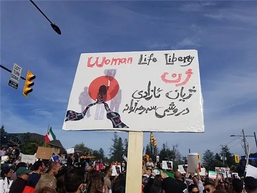 手写标语牌,上方英文“Woman Life Liberty”,下方为库尔德语文字和一个红底黑色人形剪影图案,牌子举在街头游行人群上方,背景可见交通信号灯。