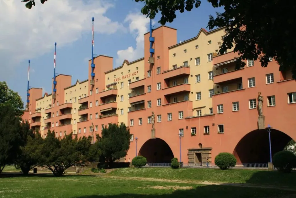 橙黄色相间的狭长住宅楼立面，多座蓝顶塔楼耸立其间，插着旗杆；楼体底层设拱门，前方是草坪与树木。
