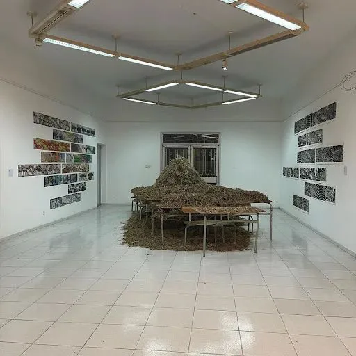 展厅中央多张课桌椅上堆满干草，两侧墙上挂着成排的黑白版画，天花板悬挂着六边形灯具组。