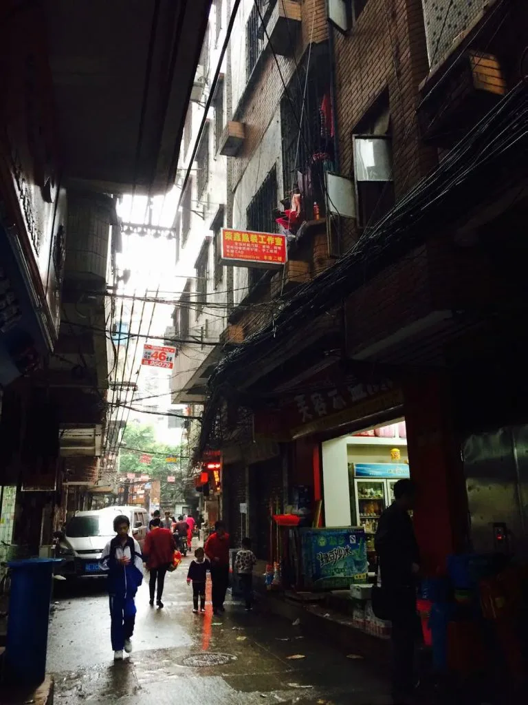 狭窄的城中村巷道两侧高楼相邻紧贴，头顶电线密布，行人和小店铺分布在潮湿的路面两侧