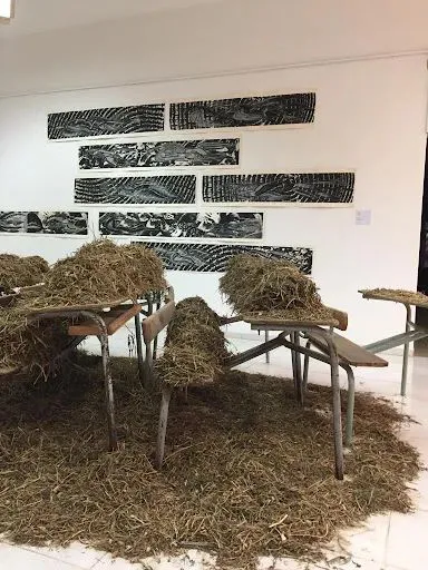 近景可见课桌椅上堆积着大量干草，背景墙上挂着数幅黑白纹理的长条状版画作品。
