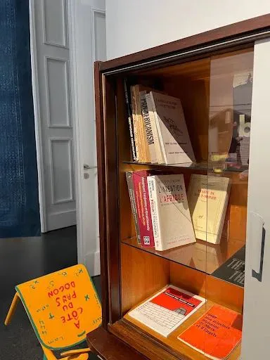 木质玻璃展柜内分层摆放多本书籍，其中可见《L'invention de l'Afrique》，柜前地上放着一把黄色折叠椅，椅面印有法文字样「A côté du pays Dogon」。