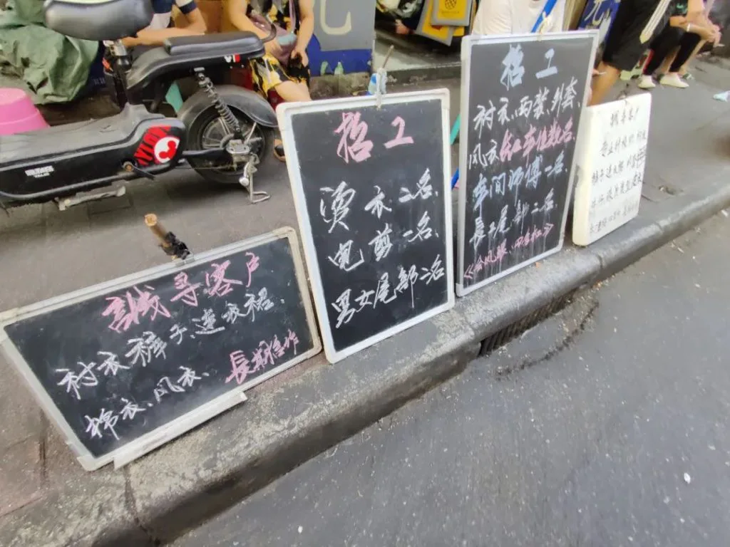 路边并排摆放着四块手写招工黑板，旁边停着一辆电动车，黑板上用彩色粉笔写着不同工种的招工信息，部分字迹潦草难以完全辨认。