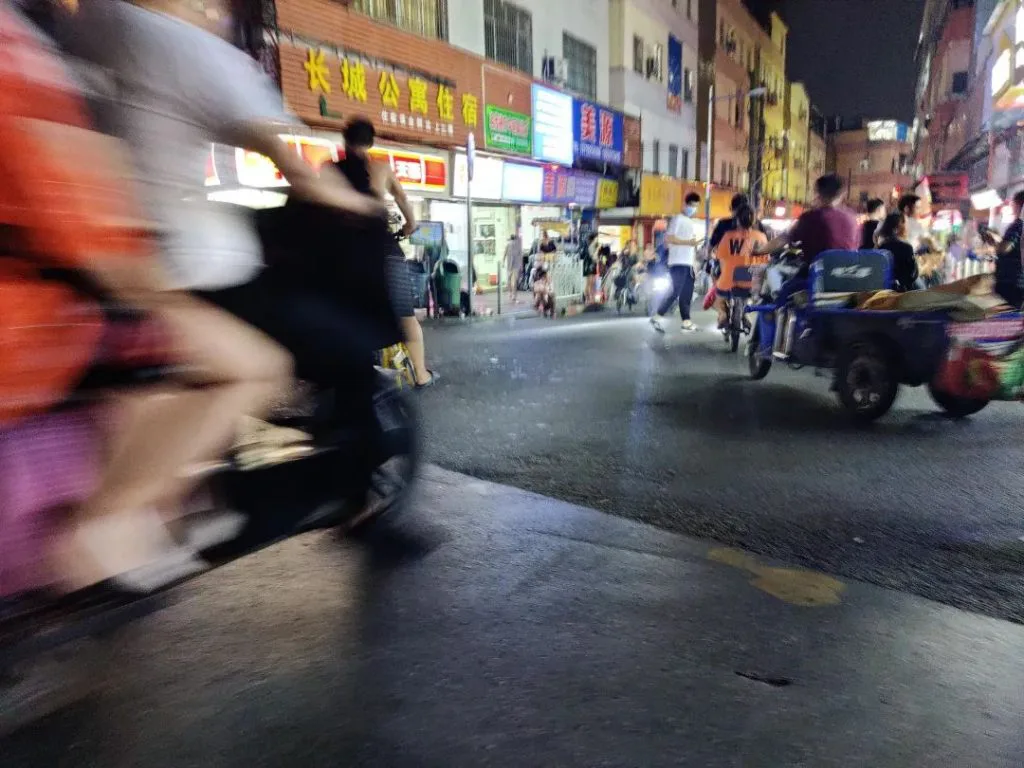 夜间街道两侧商铺灯牌林立，骑手驾驶摩托车快速穿行造成画面模糊，右侧一辆载货三轮车停靠路边，行人往来。