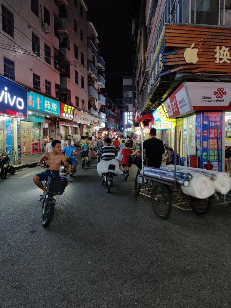 狭窄街道两旁手机店、药房等店铺灯光明亮，多名骑手驾驶摩托车穿行，右侧一辆三轮车上堆放着成捆的布匹。