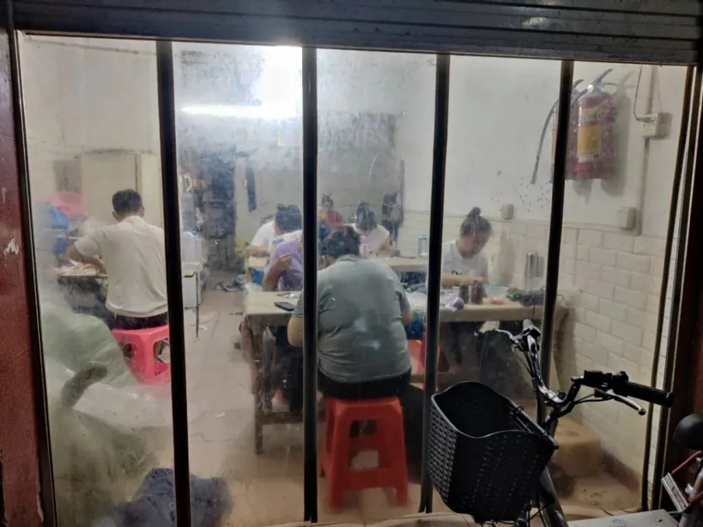 透过卷帘门可见几名工人坐在红色塑料凳上，在长桌前埋头做工，墙上挂着灭火器，门前停着一辆自行车。