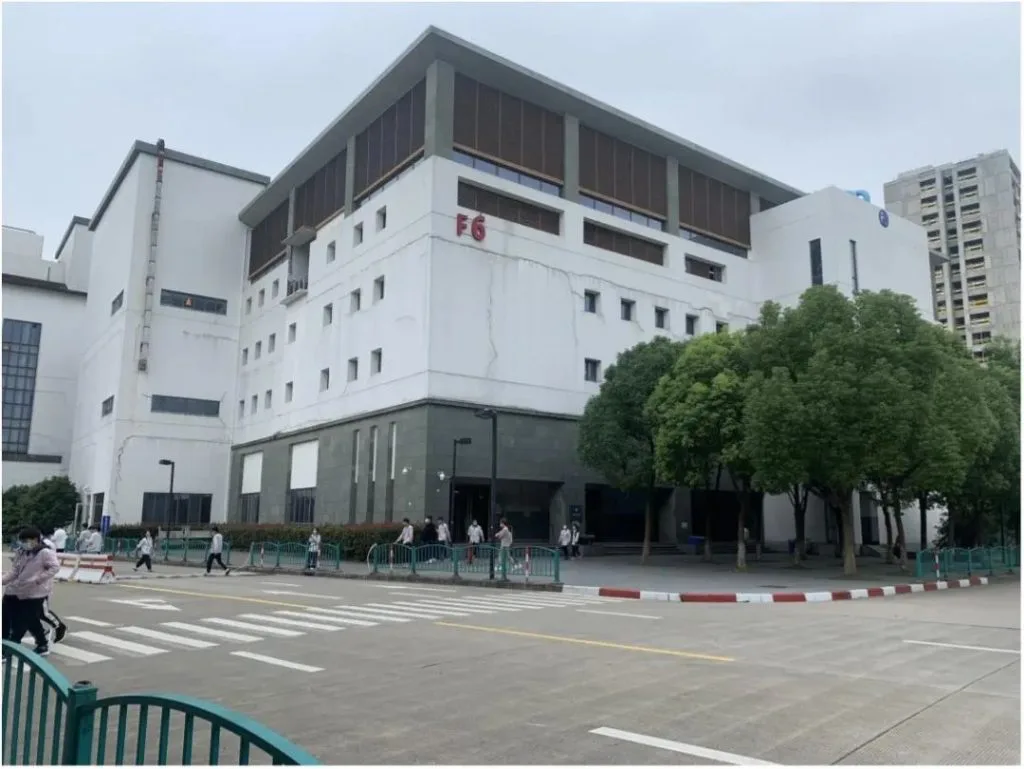 标有'F6'字样的白色多层电子厂厂房外景,几名戴口罩的员工正穿过厂区门前的马路,周围有绿化树木与护栏。