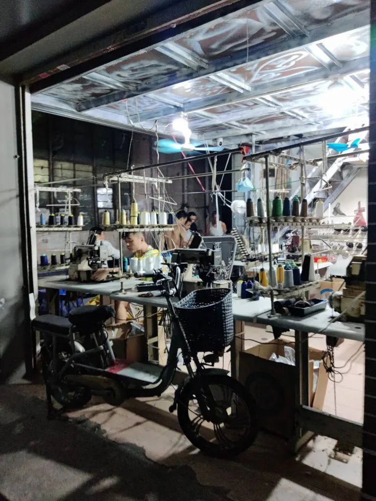 卷帘门半开的一楼作坊内，工人坐在缝纫机前工作，身后架子上挂满彩色线轴，门口停着一辆电动车。