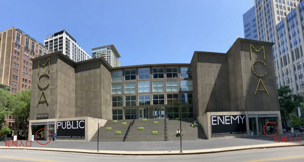 建筑正面全景,两侧塔楼贴着黄色'MCA'大字,中央宽阶梯通向玻璃门厅;图中用红圈标注两侧角落处并附文字'轮椅入口'。