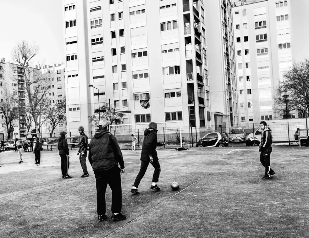 黑白照片：一群男孩在高层住宅楼下的球场上踢足球。
