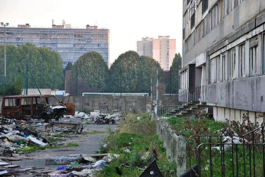 老旧的高层住宅楼下，空地上散落着大量垃圾和一辆烧毁的报废汽车，围墙上布满涂鸦。