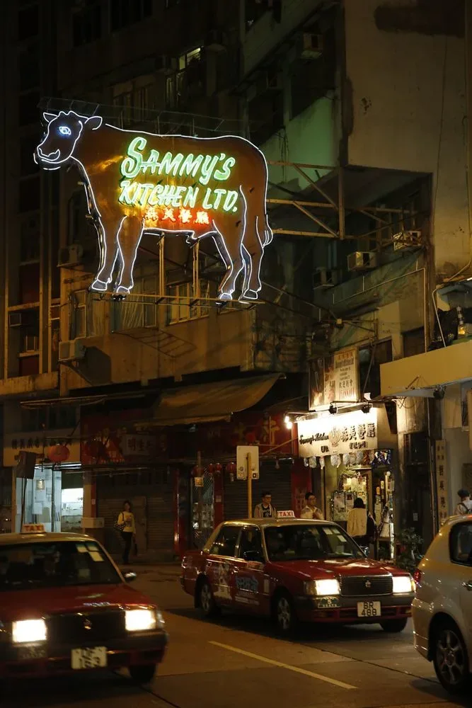 夜晚街道上方悬挂着一块巨大的牛形霓虹招牌，牛身内写着“Sammy's Kitchen Ltd.”及中文店名，下方街道有多辆红色的士驶过。