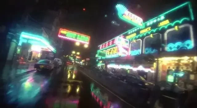 雨夜街景，两侧林立着各色霓虹招牌，湿滑地面倒映着斑斓灯光，一辆的士驶过。
