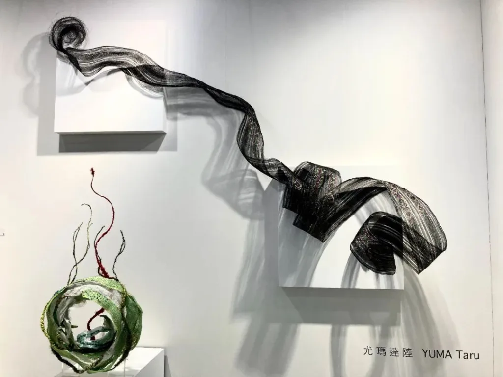 一条黑白编织长带如布匹般从墙上白色展台垂挂盘绕，下方地面摆放一件绿色环状编织物，中心探出几缕暗红色线头，墙面标注艺术家姓名尤玛·达陆。