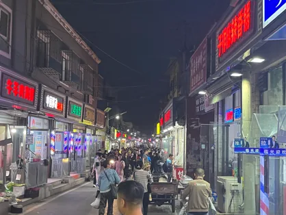 夜晚的皮村商业街灯火通明，两侧店铺霓虹招牌林立，狭窄街道上人潮涌动。