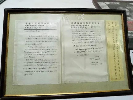 秦梅手写家书原件装裱展示，信纸抬头印着“香港致丽实业有限公司”字样，右侧贴有一张竖写的附言，具体笔迹难以辨认。