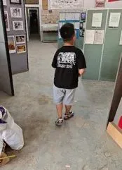 一个小男孩背对镜头站在展厅过道中,身穿印字T恤,身旁墙上贴着照片和说明牌。