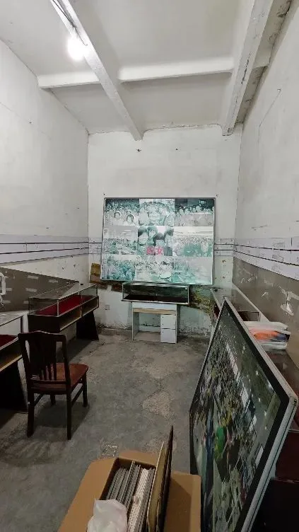 昏暗的展厅一角,墙上挂着一块由多张黑白照片拼成的九宫格展板,室内已空置,仅剩桌椅和地上的纸箱,天花板墙皮大片剥落。