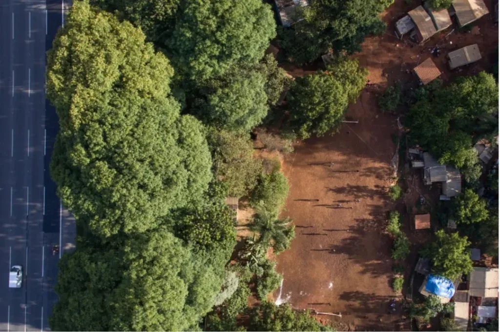 俯瞰图：画面左侧是笔直的多车道公路，右侧红土地上零星分布着简易房屋，被茂密树林环绕，公路与聚居地紧邻。