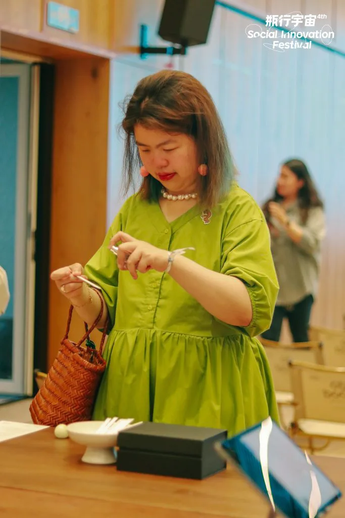 一位穿绿色连衣裙的女士站在木桌前,手拿一张纸条凑近鼻子闻嗅,桌上摆着白色小碗和黑色礼盒,背景另一位女士抱臂站立。