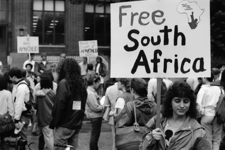 黑白照片：一名年轻女子举着写有「Free South Africa」和非洲版图图案的标语牌，背景人群中还有人举着「No More Apartheid」等标语，众人聚集在一栋建筑前。