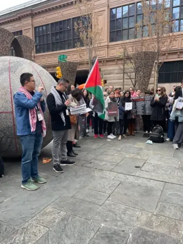 户外广场上聚集着手持标语牌的人群，一面巴勒斯坦国旗竖立其中，前方两名男子面对镜头打着手语，广场上有一座巨大的球形雕塑，背景是欧式风格建筑。