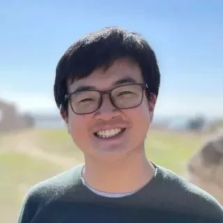 一名戴眼镜、穿深绿色上衣的男子微笑面对镜头，背景是户外虚化的草地与天空。