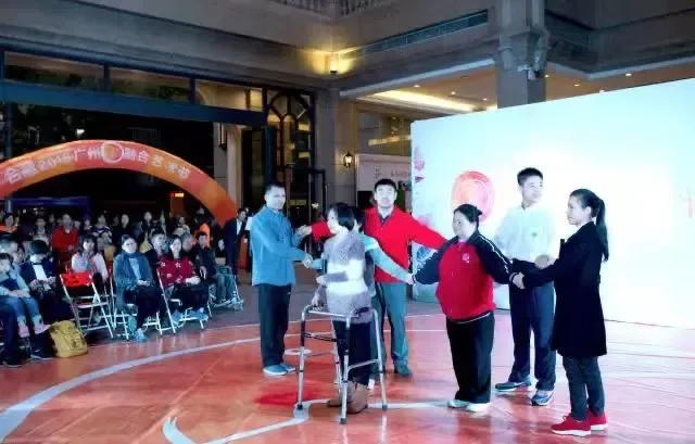 几位表演者手拉手站成一圈,其中一人扶着四脚助行器站在场地中央,身后观众席上有轮椅使用者,头顶悬挂红色拱形横幅。