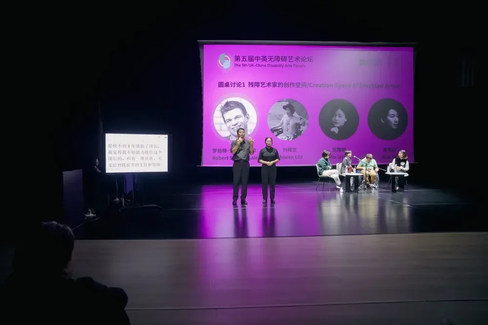 舞台大屏幕显示论坛标题与圆桌嘉宾照片,两位主持人站立讲话,右侧四位嘉宾围坐圆桌参与讨论,其中一位坐轮椅。