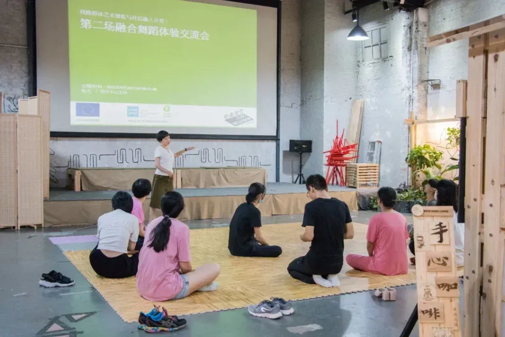 室内场地里,一人站在投影幕布前讲解,幕布上写着「第二场融合舞蹈体验交流会」,多名参与者盘腿坐在瑜伽垫上面向她。