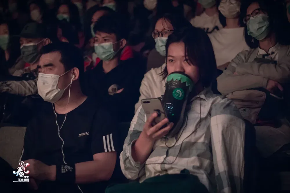 昏暗剧场中,一名女性举着一个绿色装置贴近嘴边讲述,观众席上多人佩戴口罩,部分观众戴着耳机接收讲解。