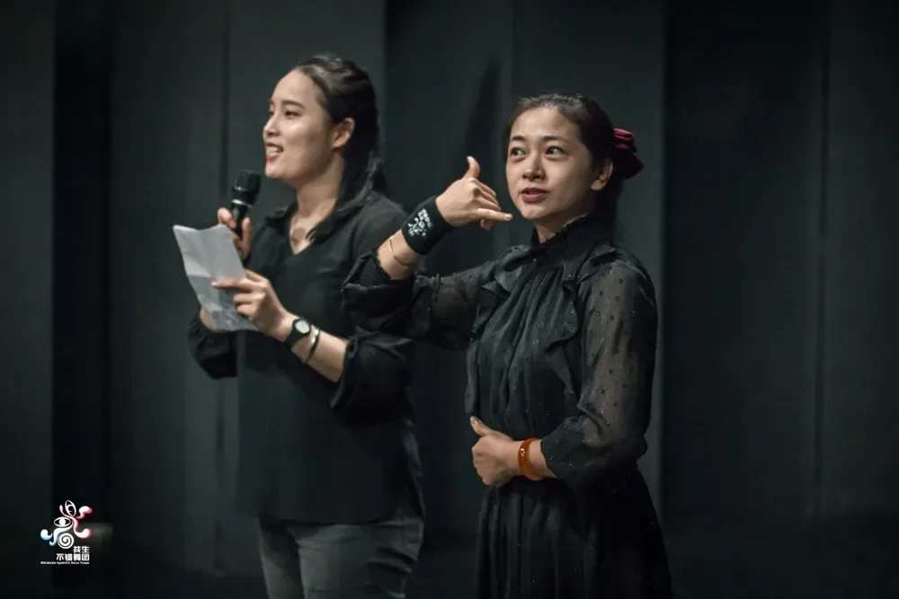 两位身穿黑色的女性站在舞台上,一人手持话筒讲话,另一人做手势打手语翻译。