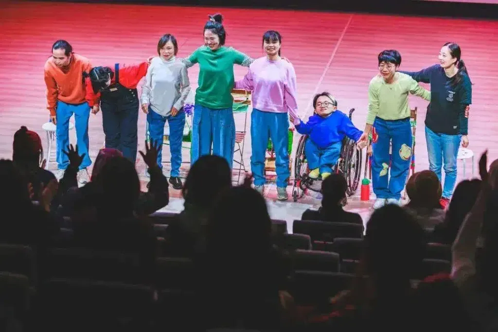 舞者们身穿彩色上衣、藏蓝色裤子站成一排谢幕,互相搭肩鞠躬,队伍中有一人坐轮椅,台下观众举手鼓掌。